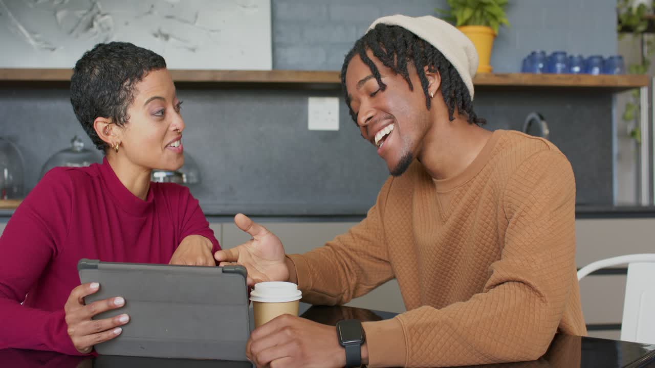 Diverse friends laughing at tablet content while tapping screen and passing coffee cup at cafe