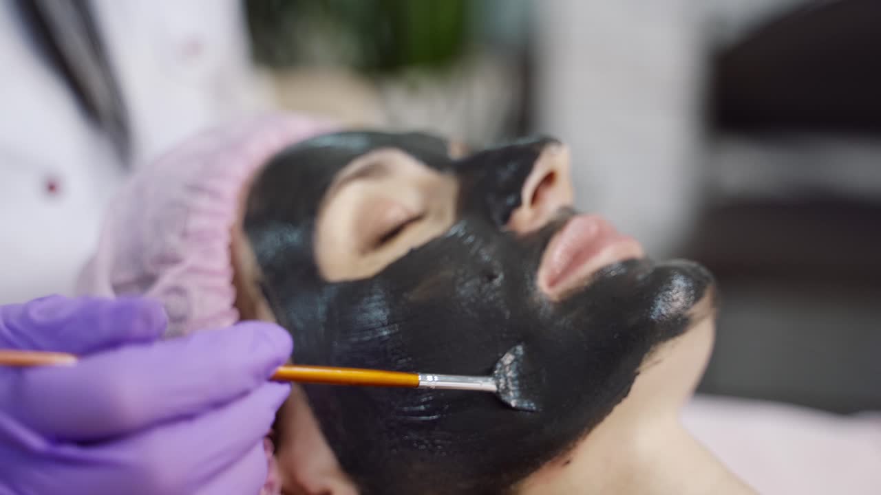 A specialist is spreading mask with brush on the face of the woman in the spa salon. Beauty industry. Close-up. Blurred background.