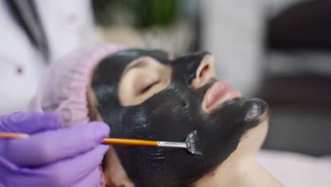 a specialist is spreading mask with brush on the face of the woman in the spa salon. beauty industry. close-up. blurred background.