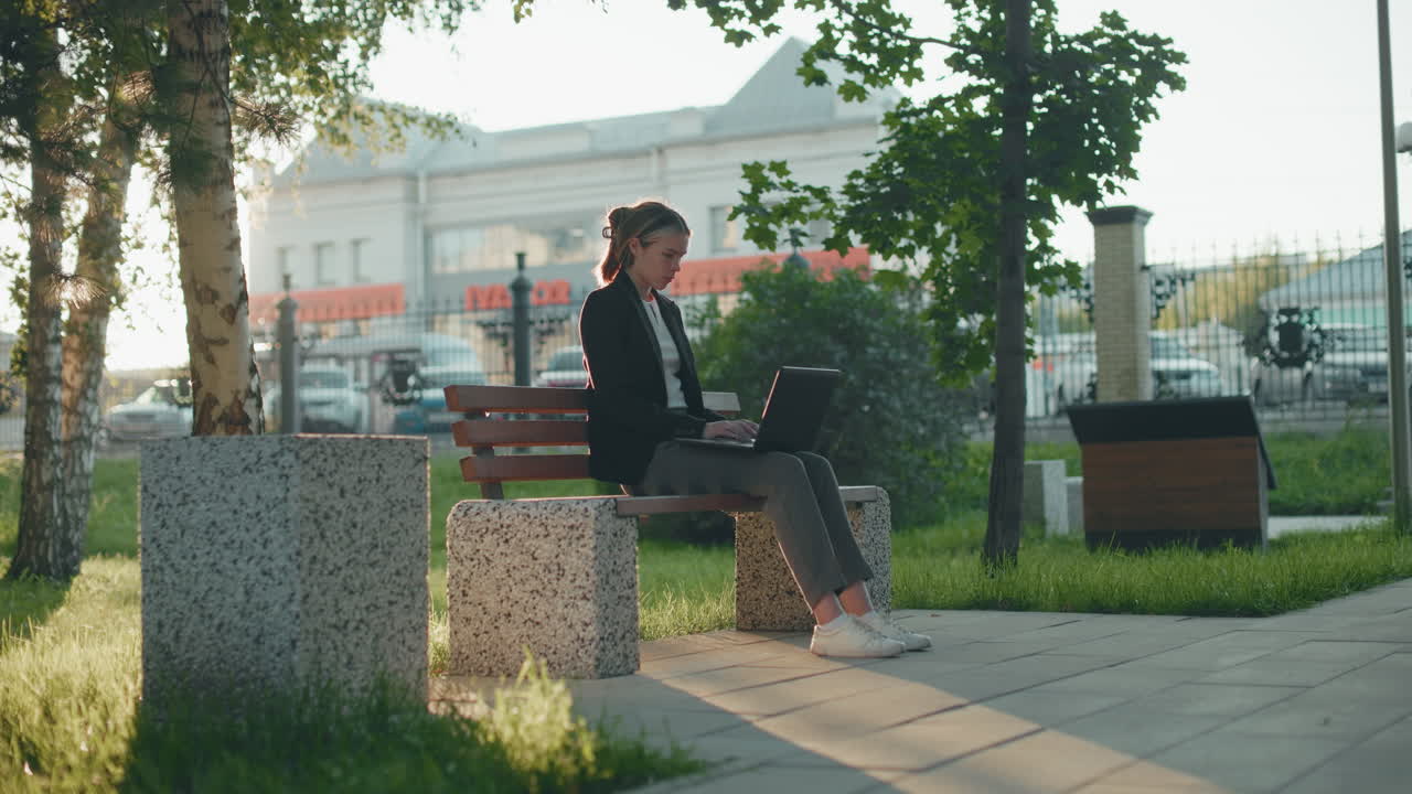 L'effetto silhouette mette in risalto una donna che lavora all'aperto in un parco mentre sorseggia un succo da una tazza con un laptop sulle gambe, circondata da vegetazione, auto parcheggiate e un autobus in movimento visibile sullo sfondo in lontananza