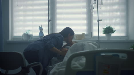 mother kissing sick daughter lying in bed. supportive parent visit patient ward