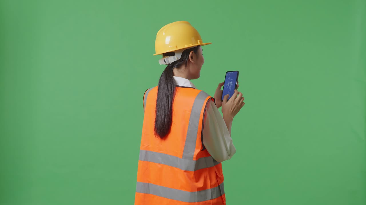 안전 헬을 입은 아시아 여성 엔지니어가 녹색 화면 배경 스튜디오에 서 있는 동안 스마트폰에서 풍력 터빈 블루프린트를 보고 있는 뒷면