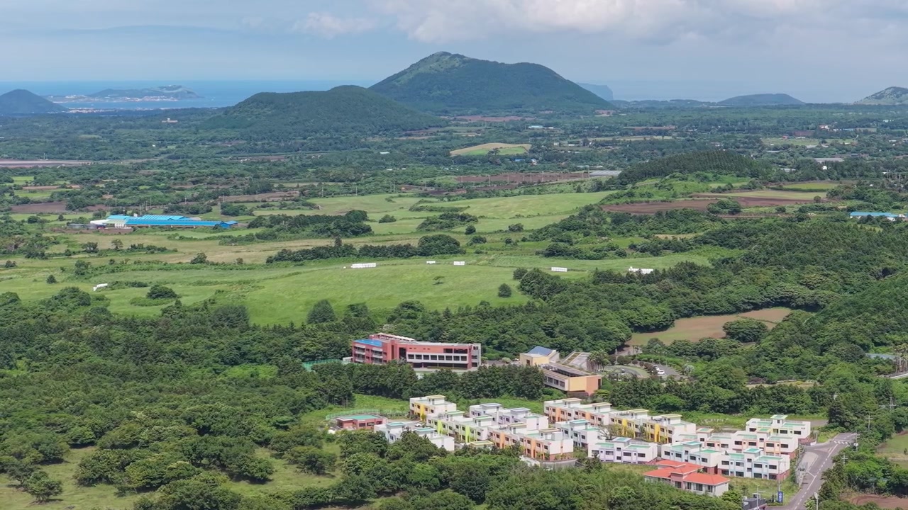 Vista aérea horizontal del humedal Dongbaek Dongsan en el este de la Isla de Jeju, Corea del Sur, mostrando edificios, campos verdes, bosque y una montaña volcánica mientras la cámara se desplaza lentamente hacia la izquierda.