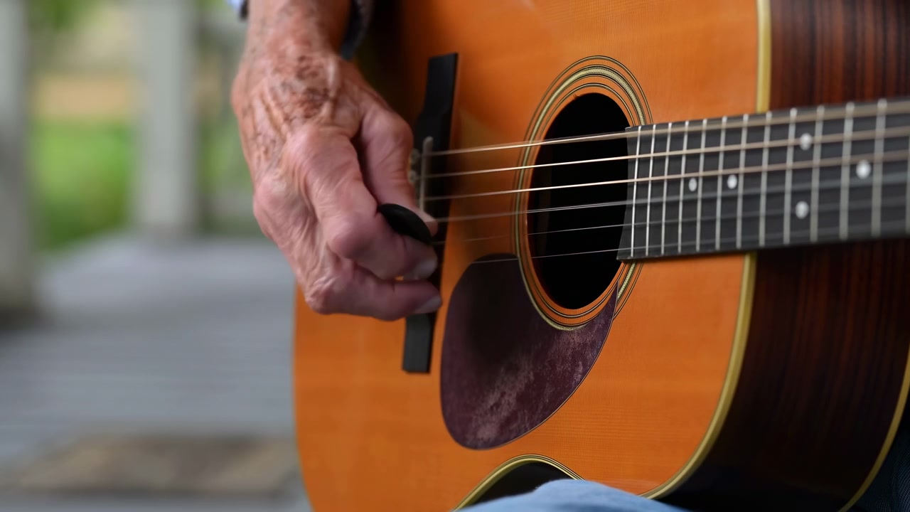 Vista ravvicinata di una mano che si muove sulle corde di una chitarra acustica, con la calda texture del legno e la buca di risonanza visibili, e un morbido sfondo esterno che crea una composizione naturale e calma.