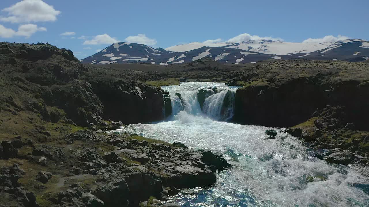 Flying a drone up river with pure and cold water towards a waterfall in the highlands of Iceland. Snow capped mountains in the background.