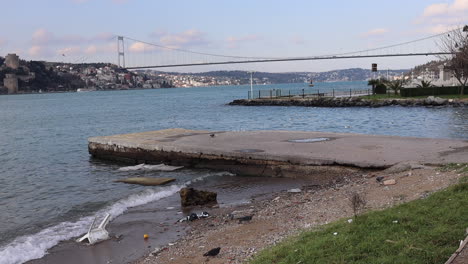 Waste-And-Plastic-Garbage-Washed-Ashore-On-Picnic-Ground-With-Fatih-Sultan-Mehmet-Bridge-In-The-Distance-In-Istanbul,-Turkey