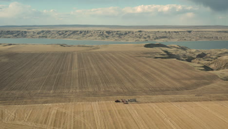 seeding tractor working at the vast farmland with calm lake at background in - aerial wide shot