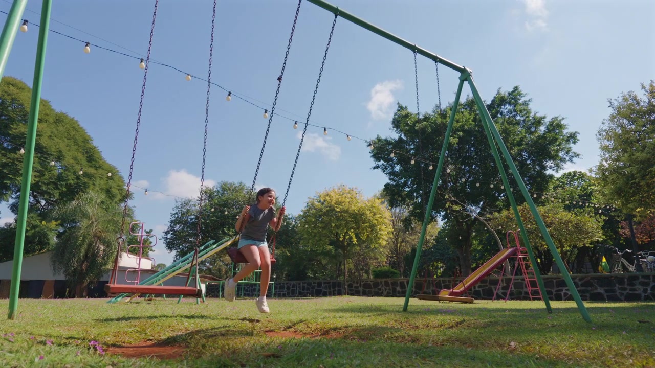 Una niña se columpia alegremente en un parque bajo un cielo azul claro, rodeada de equipos de juegos infantiles. Ideal para temas de familia, ocio o actividades al aire libre.
