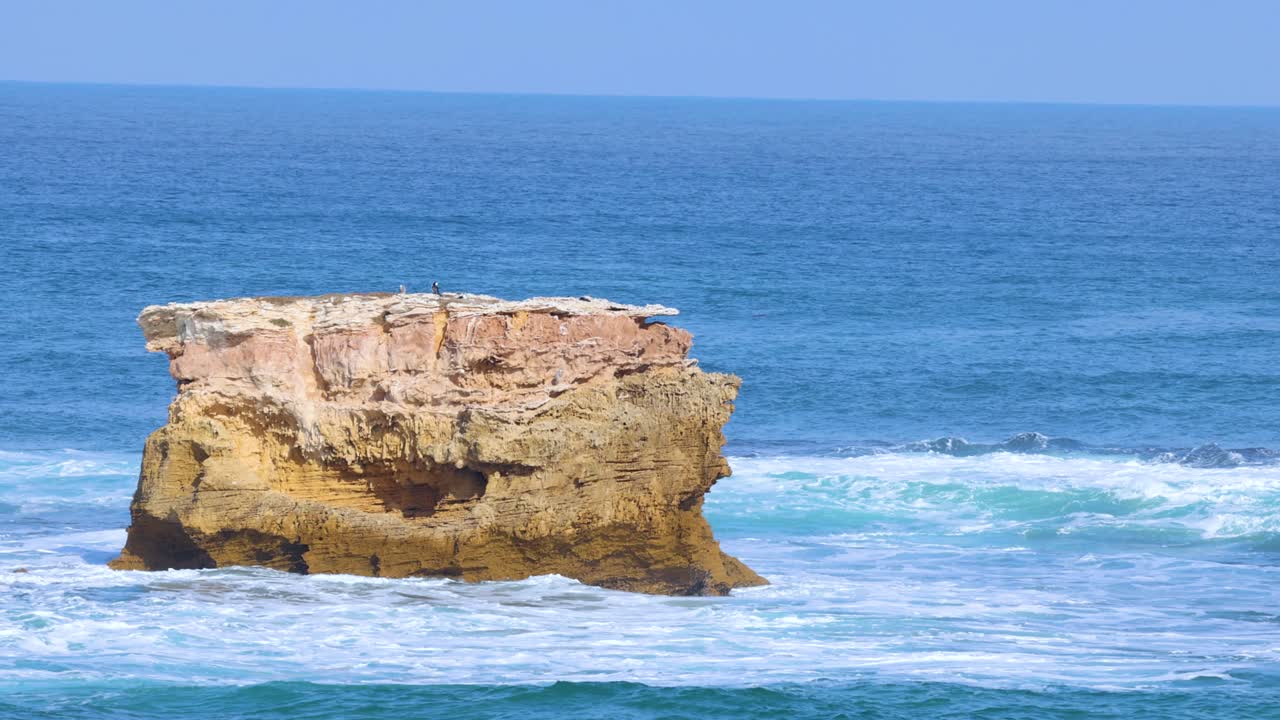 Ocean waves crash against a solitary rock formation on Mornington Peninsula, under bright daylight, showcasing nature's power and beauty