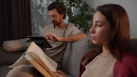 Close-up-view-of-a-young-brunette-girl-in-a-red-sweater-reading-a-book-while-her-brunette-boyfriend-with-stubble-turns-his-attention-to-her-on-the-sofa-in-a-modern-apartment-at-home