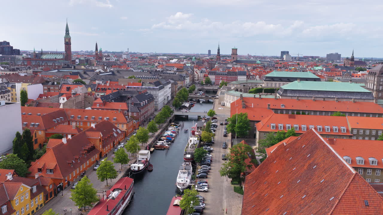 Aerial drone view of historic Christianshavn canal lined with boats and red-roofed buildings, with Copenhagen's skyline in the distance in Denmark