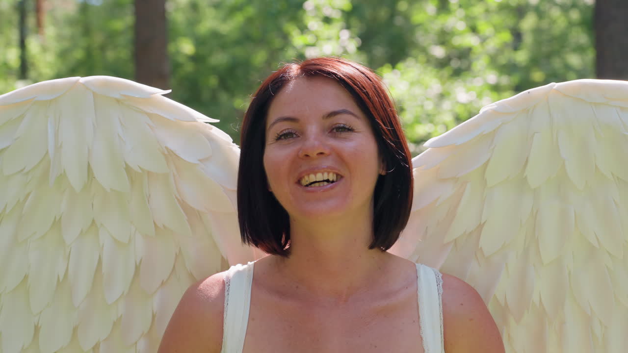 Female model dressed like angel in forest tilts head and smiles warmly, wings glowing in sunlit greenery, gentle breeze stirring feathers while smile softens and fades