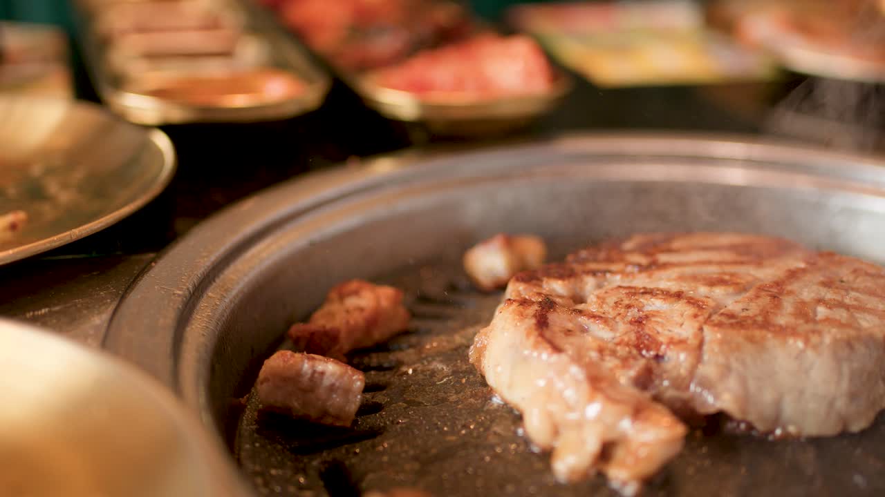 Tongs flip pork steak on tabletop grill, warm lighting, shallow depth, Korean barbecue restaurant setting