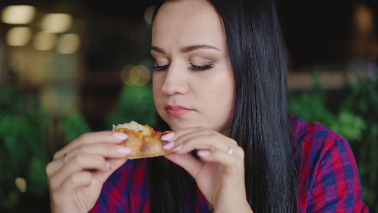 beautiful brunette in a plaid shirt is eating pizza with enjoy in the cafe on the background of the interior
