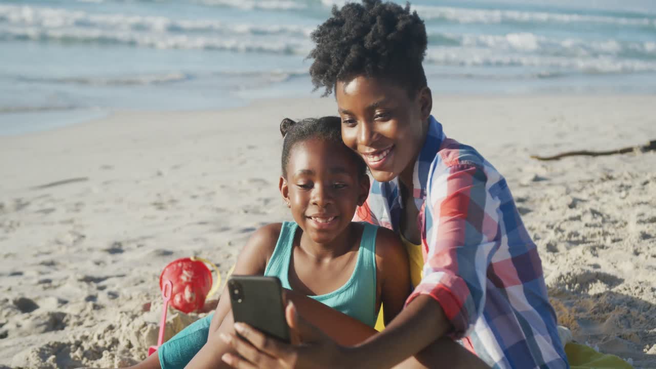 행복한 아프리카계 미국인 어머니가 해변에서 딸과 셀카를 찍고 있습니다.
