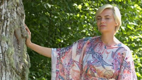 una mujer vestida de color parada sola con una de sus manos tocando el árbol meditando