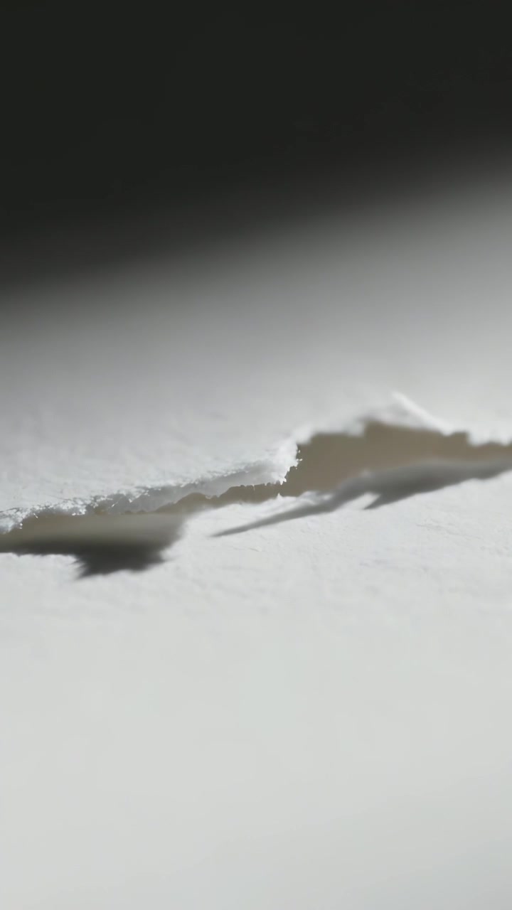 Vertical video: Shifting light casting shadow across torn white paper edge on desk, showing fibers