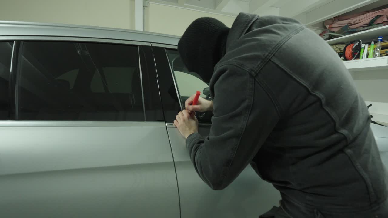A masked intruder tries to break into a parked car inside a garage, portraying car theft risk, crime, and the need for vehicle security and insurance