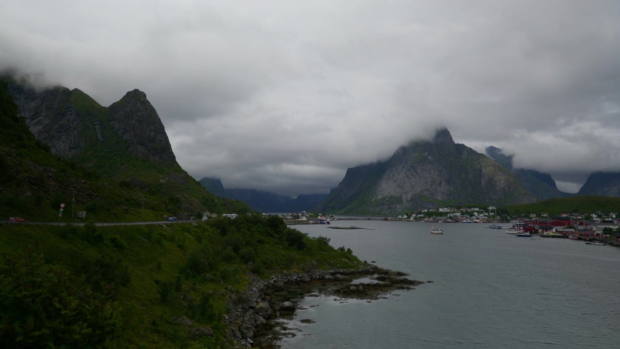 Stille fjordvand løber forbi en kystvej og en lille landsby under lave sommerskyer på Lofoten i Norge