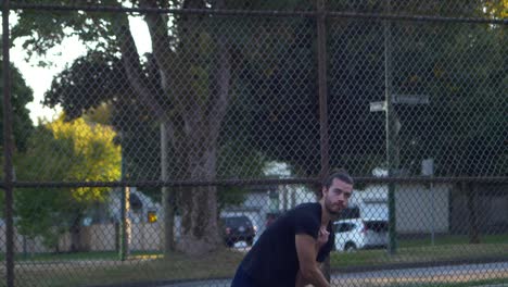 hombre con barba y bollo de hombre en un parque sirve una pelota de tenis con cerca de cadena en segundo plano