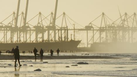 people silhouettes walk on beach in sunset with industrial port crane backdrop