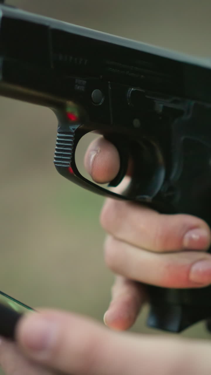 una vista en primer plano de un hombre con una chaqueta verde sosteniendo un teléfono celular y un arma, con su dedo en el gatillo, usando uno de sus dedos para operar su teléfono