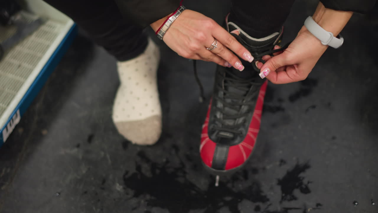 Close view lady lacing red black ice skates while sitting on bench wearing black pants and white dotted socks, focusing on careful preparation before skating on icy rink with shoelaces pulled tight by hands