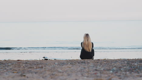 Rear-View-Of-Woman-Wearing-Wetsuit-Sitting-On-Surfboard-And-Looking-Out-To-Sea