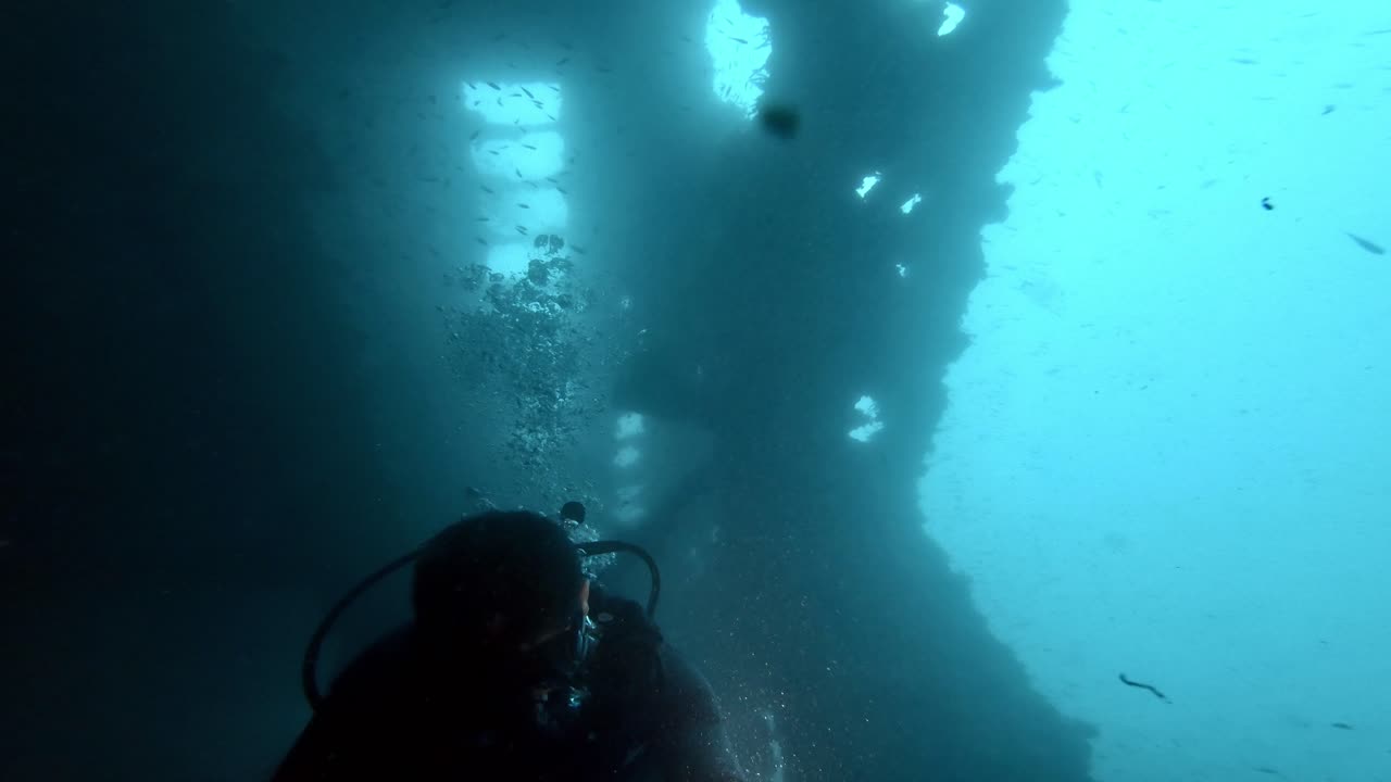 깊은 푸른 물에서 오래된 민간 난파선 내부로 잠수하는 스쿠버 다이버, 표면에서 오는 햇빛으로 어두운 풍경이 밝아집니다.