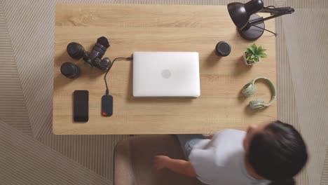 top view of asian woman color grading walking into the workspace sitting down and using a laptop next to the camera editing the video at home