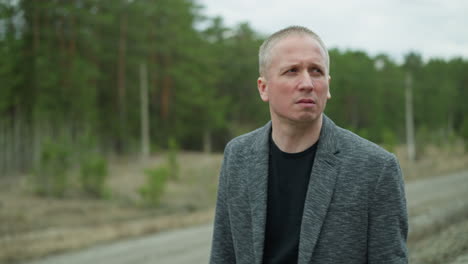 a close-up side profile of a middle-aged man with short hair, wearing a grey suit jacket and a black shirt, walking outdoors with a blurred forest background