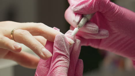 close-up of nail technician in pink gloves using electric file to shape customer nail as fine dust scatters. client wears gold ring while hand is gently held for precision nail care and grooming