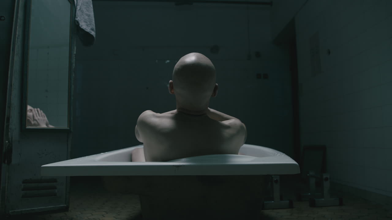 Pale bald skinny man takes a bath in the creepy old bathroom at night