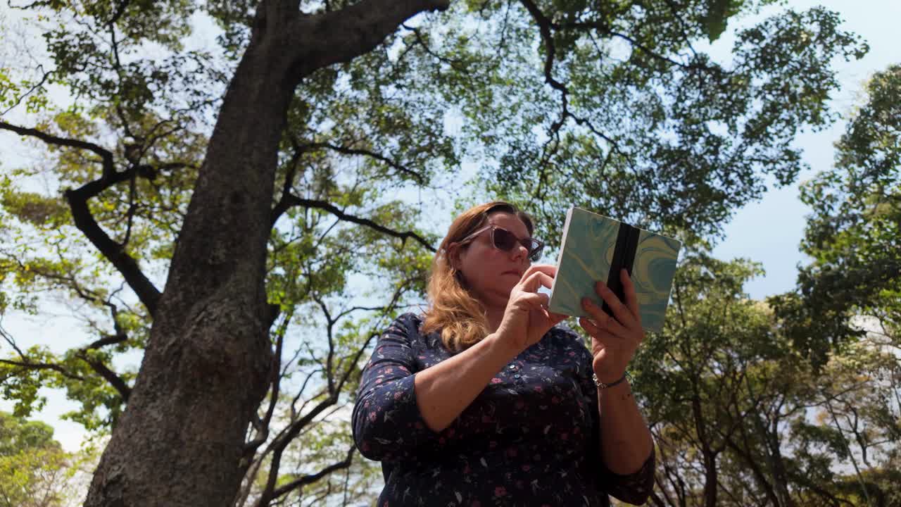 Woman relaxing and writing in a notepad under a tall green tree, enjoying a peaceful mindfulness moment