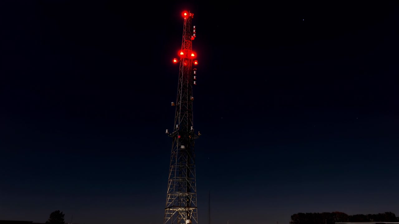 Vista notturna dal basso verso l'alto di un'alta torre di comunicazione metallica con luci di segnalazione rosse brillanti, deboli sagome di alberi e tetti vicino all'orizzonte, cielo scuro profondo, forte contrasto e un elemento tecnico isolato.