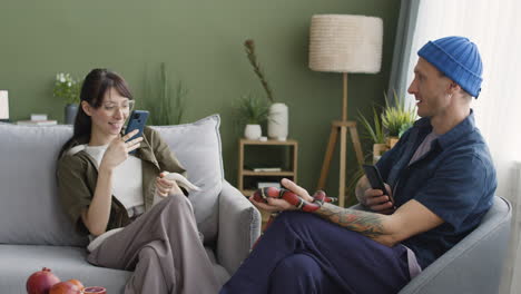 young woman and tattoed man holding their pet snakes and taking photo with phone while sitting on sofa at home