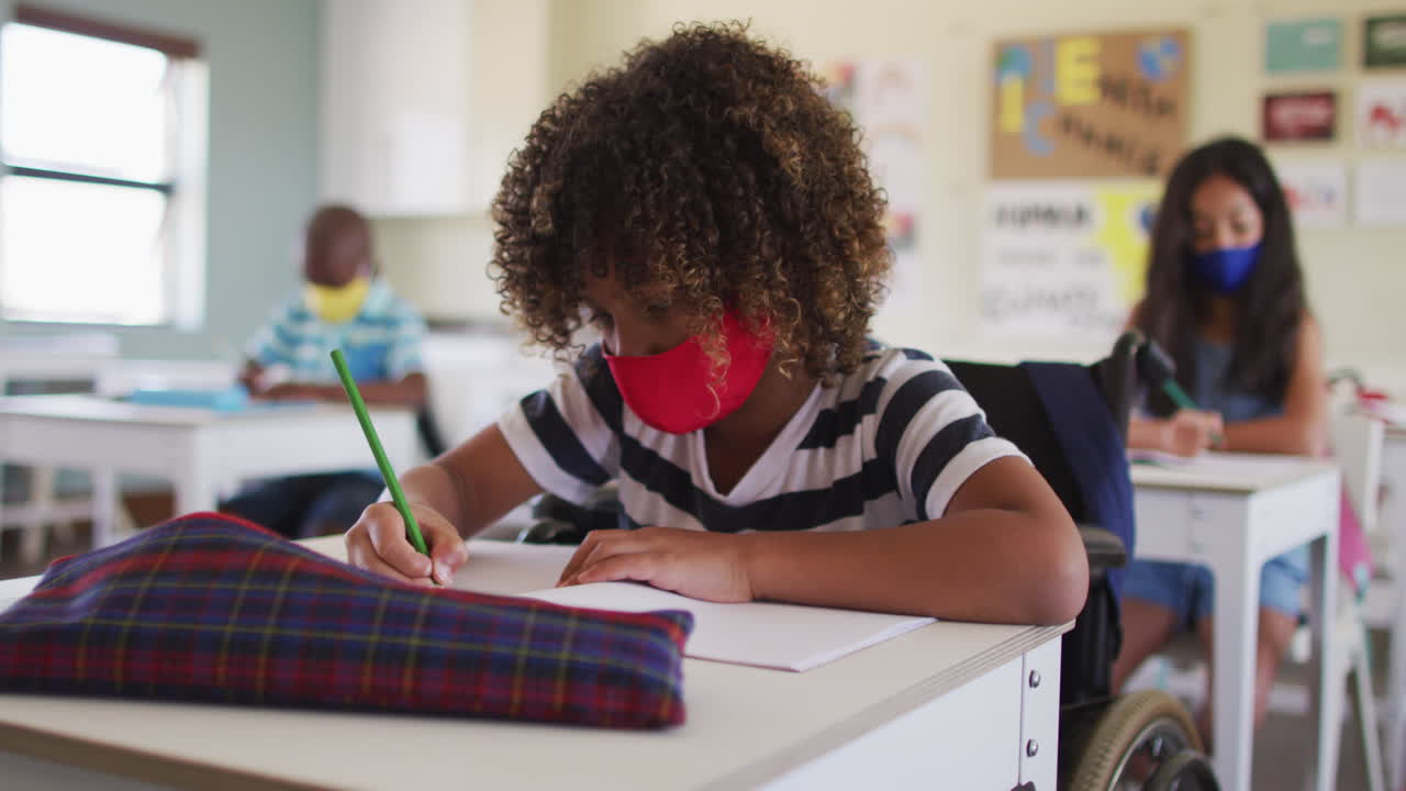 Disable boy wearing face mask writing while sitting on his wheelchair in class