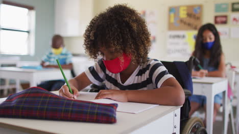 Inhabilitar-A-Un-Niño-Que-Usa-Mascarilla-Escribiendo-Mientras-Está-Sentado-En-Su-Silla-De-Ruedas-En-Clase