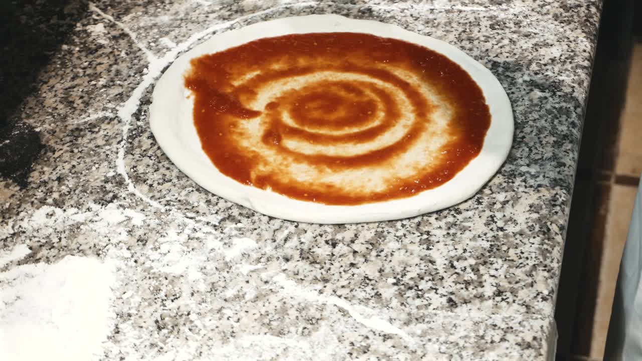 Footage of chef pouring tomatoes sauce on pizza dough.