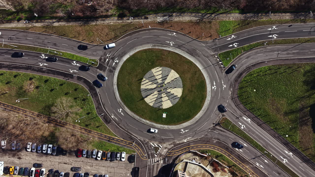 Cars move around a roundabout near a parking lot with green grass and pathways in the area during daylight hours