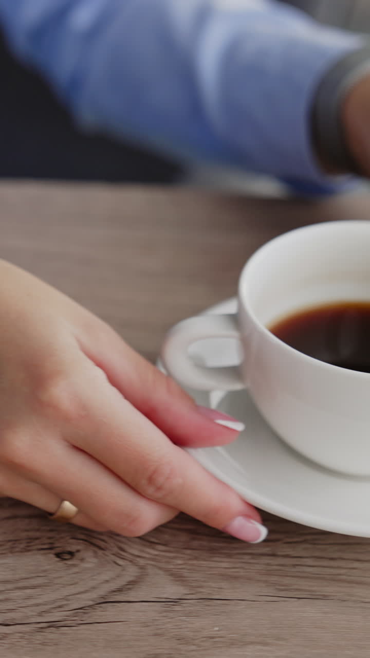 White cup of black coffee on the wooden table. Female hands pull the cup closer. Laptop and phone are on the desk beside. Close up. Vertical video