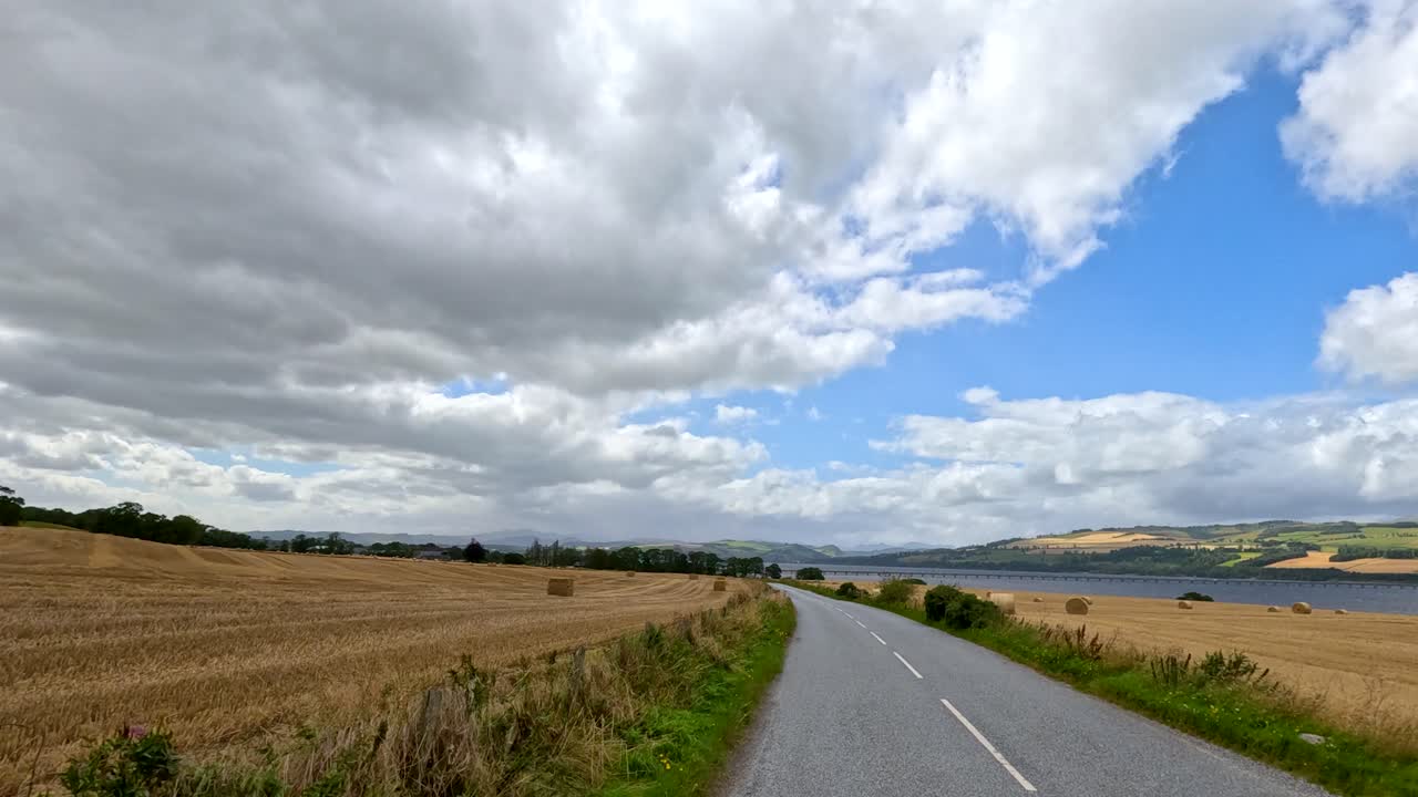 A moving vehicle travels down a winding country road bordered by harvested hay fields under dramatic, partly cloudy skies in the Scottish countryside