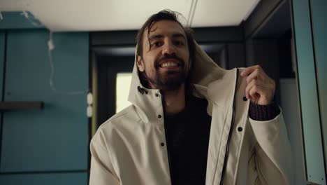 Portrait-of-a-happy-brunette-guy-who-takes-off-his-white-jacket-after-a-walk,-looks-at-the-camera-and-smiles.-Video-filmed-in-high-quality