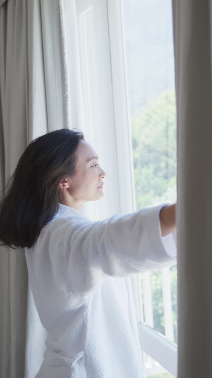 Vertical video of happy asian woman opening curtains in bedroom, in slow motion