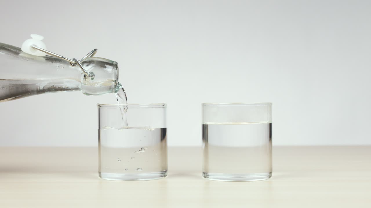 Clear water poured from bottle into glass beside another, bright lighting, static camera, neutral background