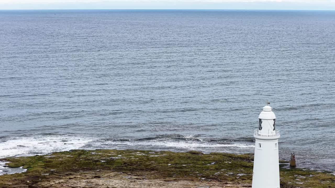 Drone camera smoothly flies over a white lighthouse and red-roofed buildings on a rocky shoreline, revealing the expansive ocean under soft daylight