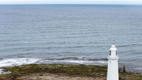 drone camera smoothly flies over a white lighthouse and red-roofed buildings on a rocky shoreline, revealing the expansive ocean under soft daylight