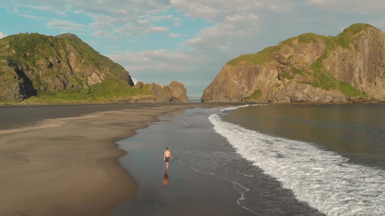 Aerial drone shot of young man walking on black sand vulanic Whatipu Beach by ocean towards incredible rock formations