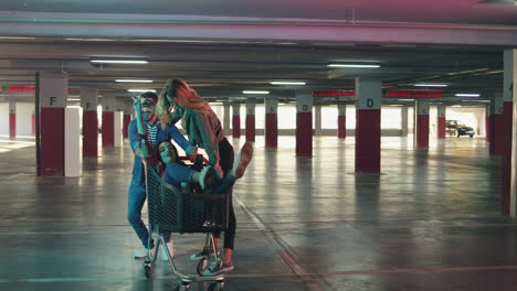 young cheerful caucasian man having fun with two girls while carrying them in the trolley in a empty parking