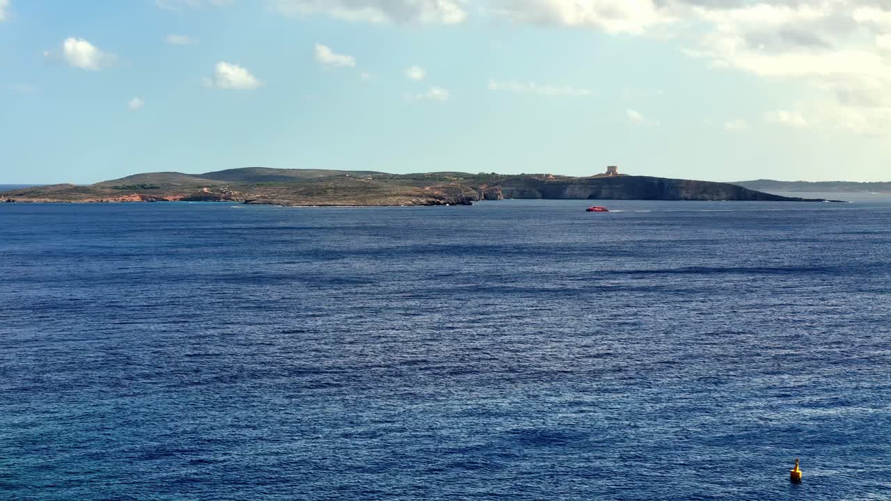 Vista panoramica grandangolare dell'isola di Comino sotto un sole estivo splendente, con la Torre di Santa Maria che si affaccia sulle acque blu intenso del Mar Mediterraneo, cielo limpido e nuvole soffici durante una calda giornata a Malta